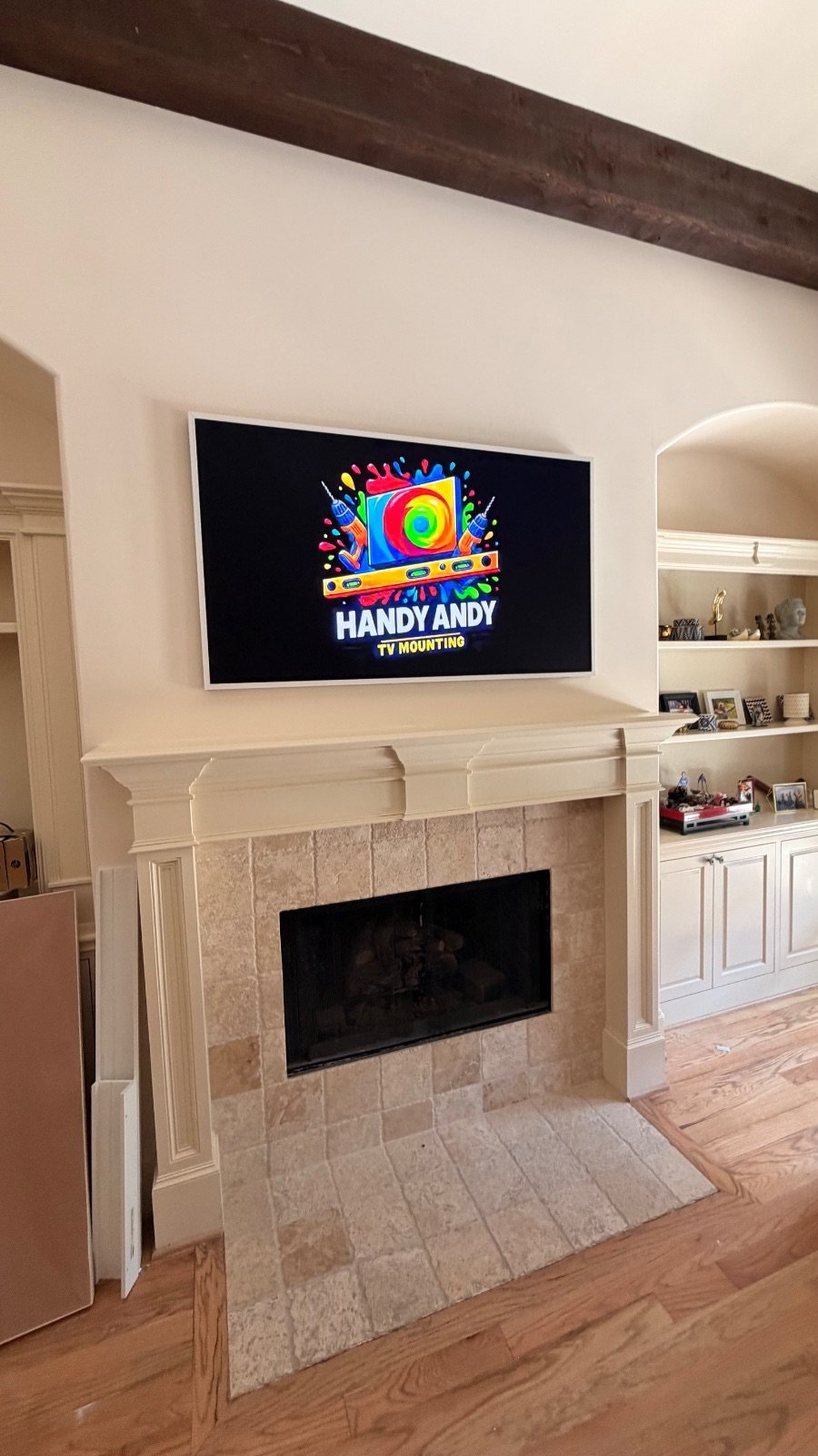 Flat screen TV mounted above a travertine fireplace displaying a Handy Andy logo, with built-in shelving visible to the right
