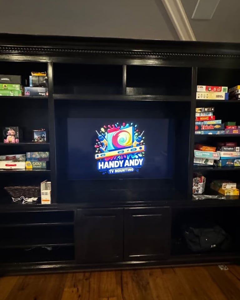 Black entertainment center with TV displaying Handy Andy TV Mounting logo on the screen, surrounded by shelves of collectibles and games