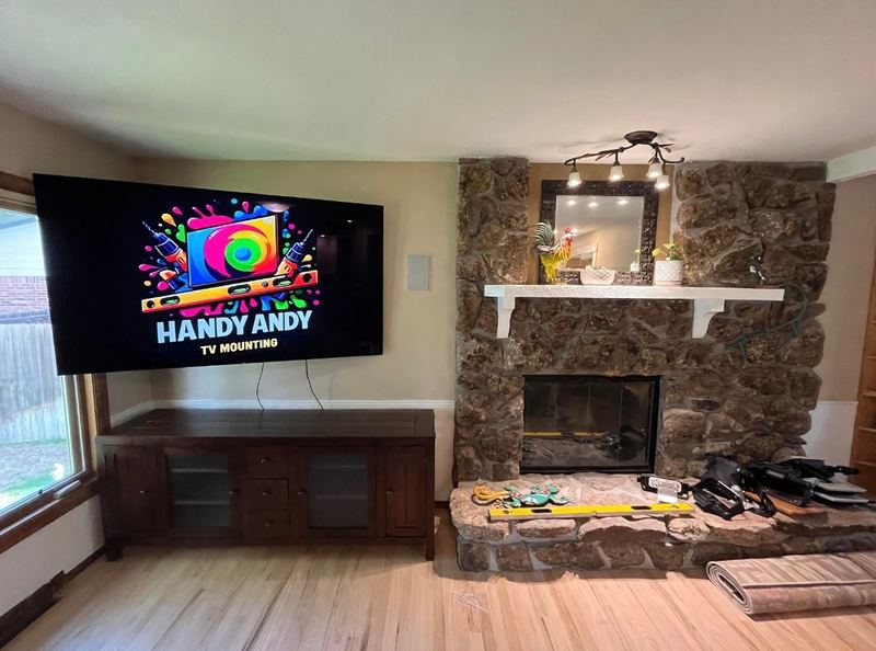 Large TV mounted on a beige wall above a wood TV stand, next to a rock fireplace with mounting tools on the hearth