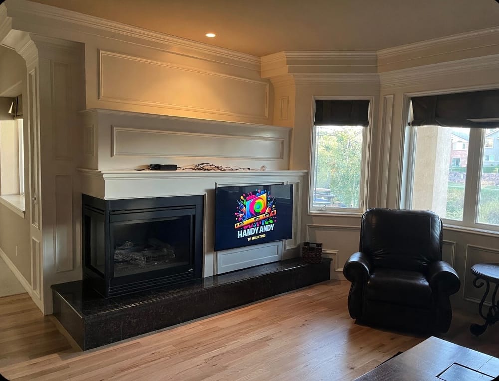 TV mounted above a white fireplace with a black hearth and recessed paneling in a living room, displaying the Handy Andy logo