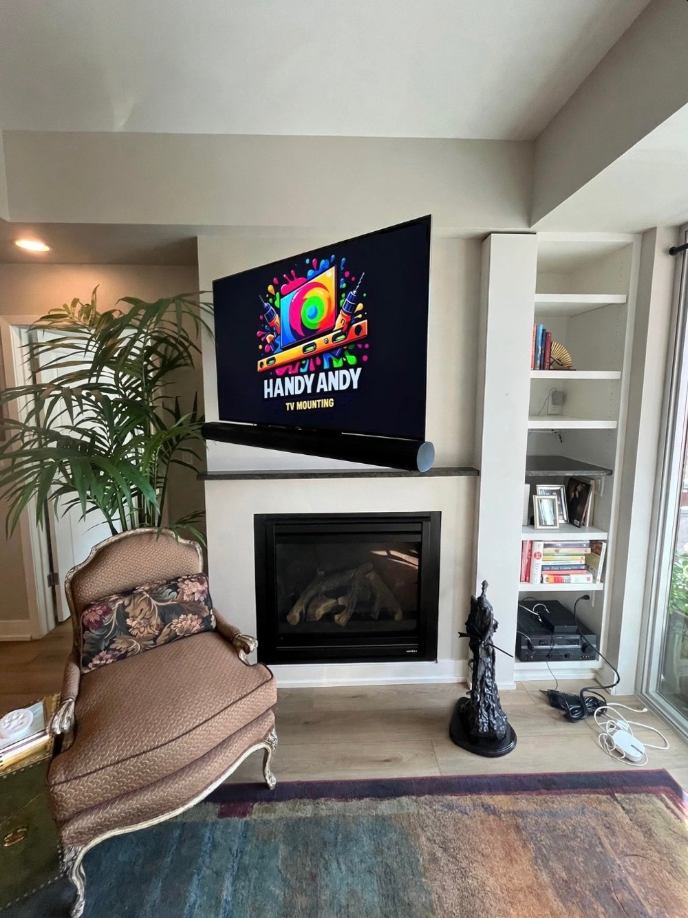 TV mounted above a gas fireplace between white built-in shelves with a tall plant and armchair, showing the Handy Andy logo