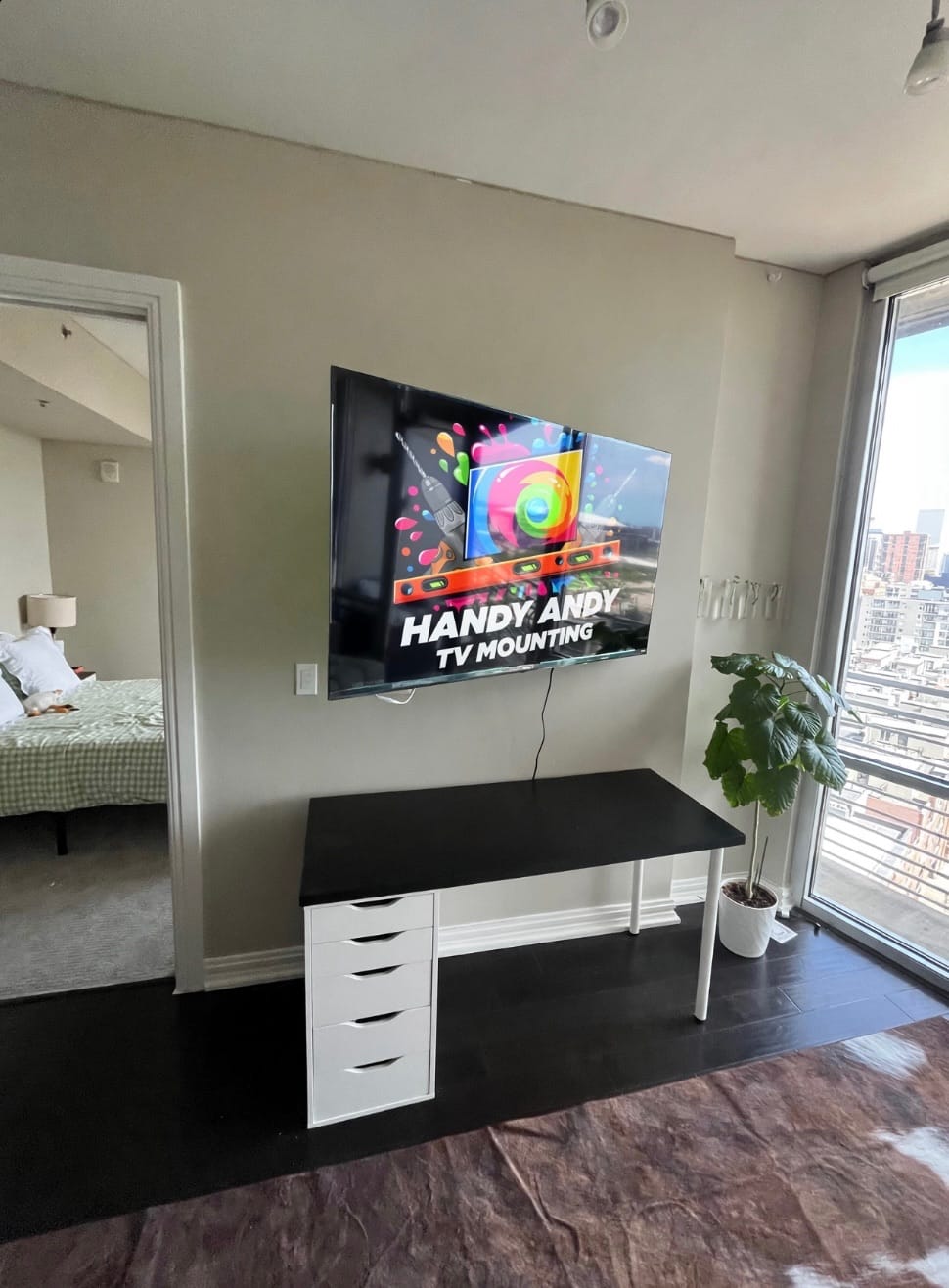 TV mounted on a wall above a black desk with a plant beside a bedroom window, displaying the Handy Andy logo