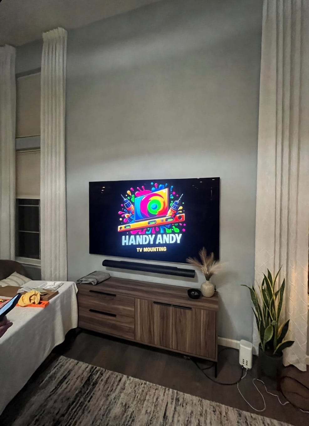 TV mounted on a gray wall beside tall white columns above a wood console with a soundbar, displaying the Handy Andy logo