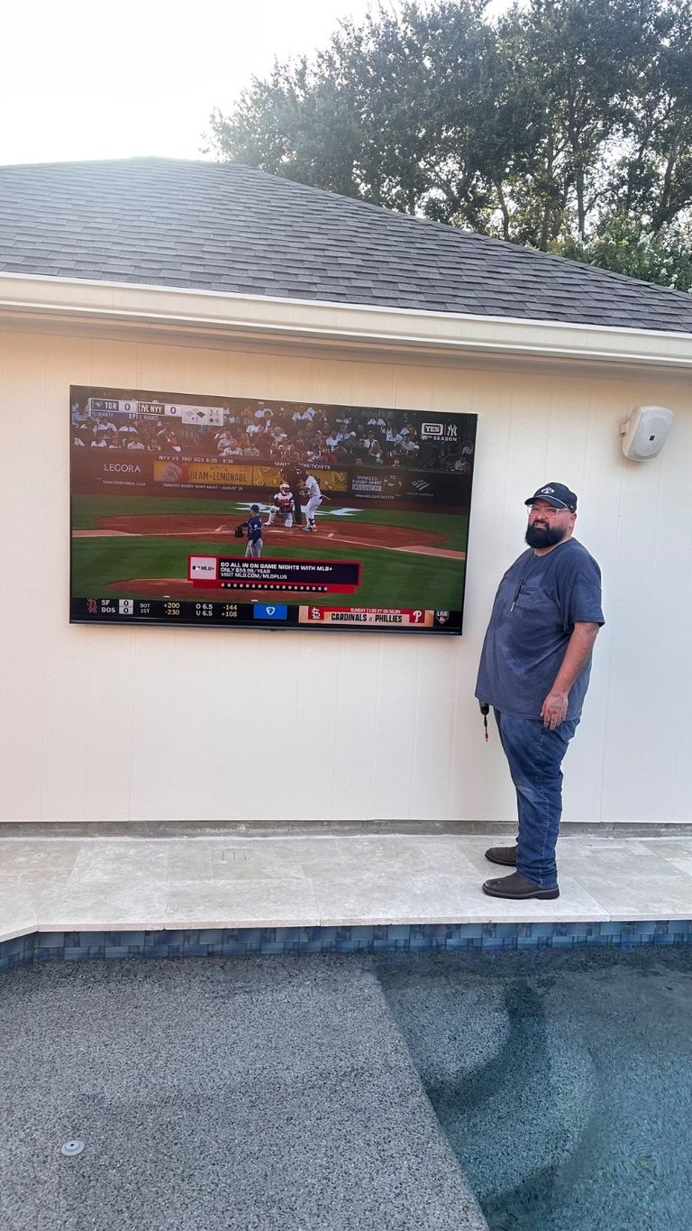 Man in blue shirt and cap standing by a pool, watching a baseball game on an outdoor TV mounted on a white wall