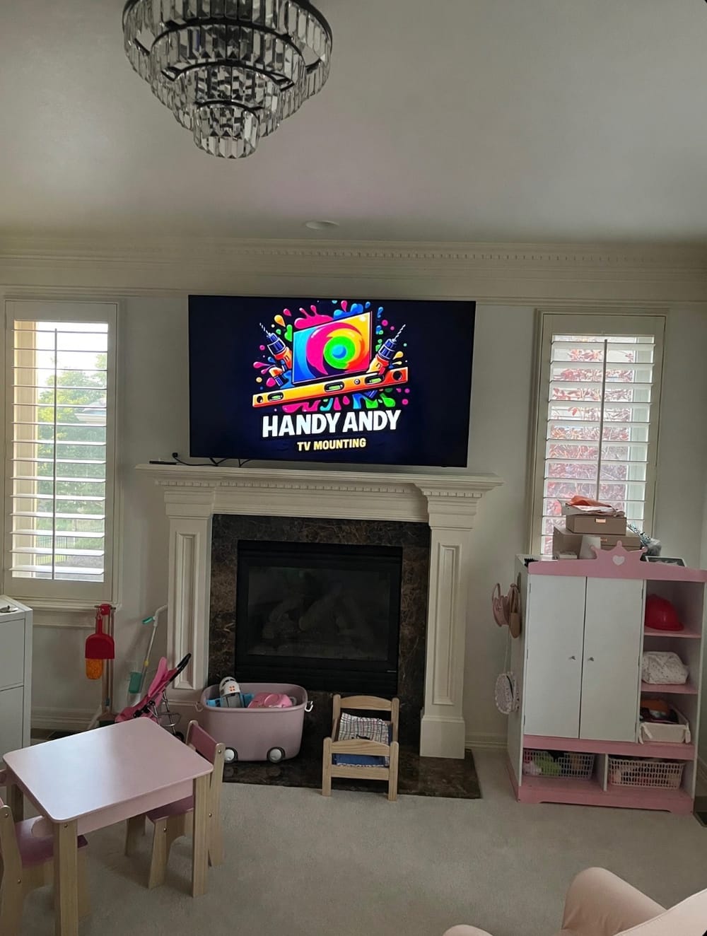 TV mounted above a white fireplace with a crystal chandelier overhead, displaying the Handy Andy logo, in a family room with a play area