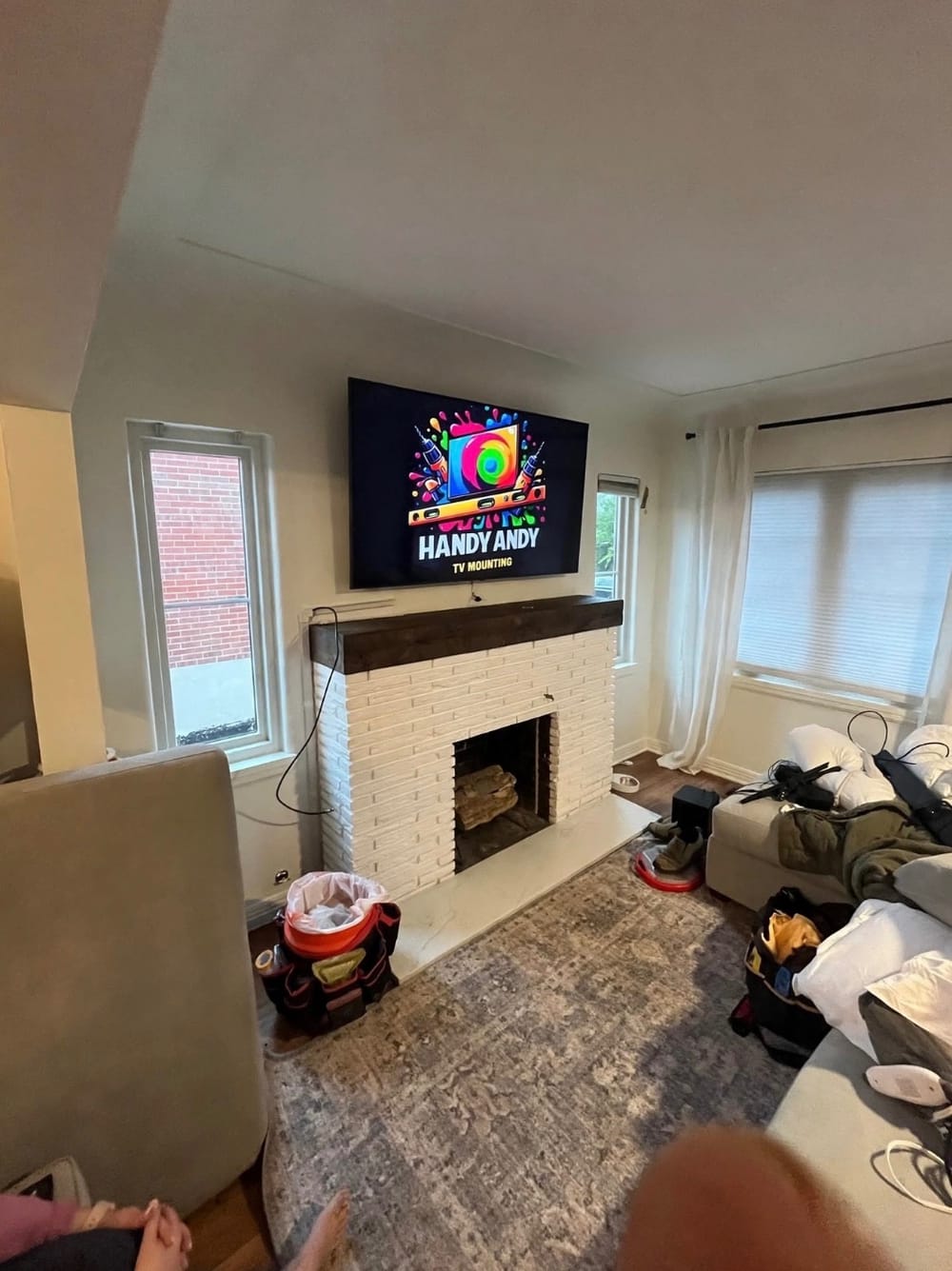TV mounted above a white painted brick fireplace with a wood mantel, displaying the Handy Andy logo