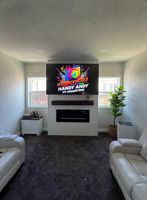 Modern living room with wall-mounted TV displaying Handy Andy TV Mounting logo, fireplace below, two white chairs, potted plant, and windows on either side