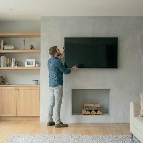 Man in blue plaid shirt reaching toward wall-mounted TV in modern living room with fireplace and wooden shelving