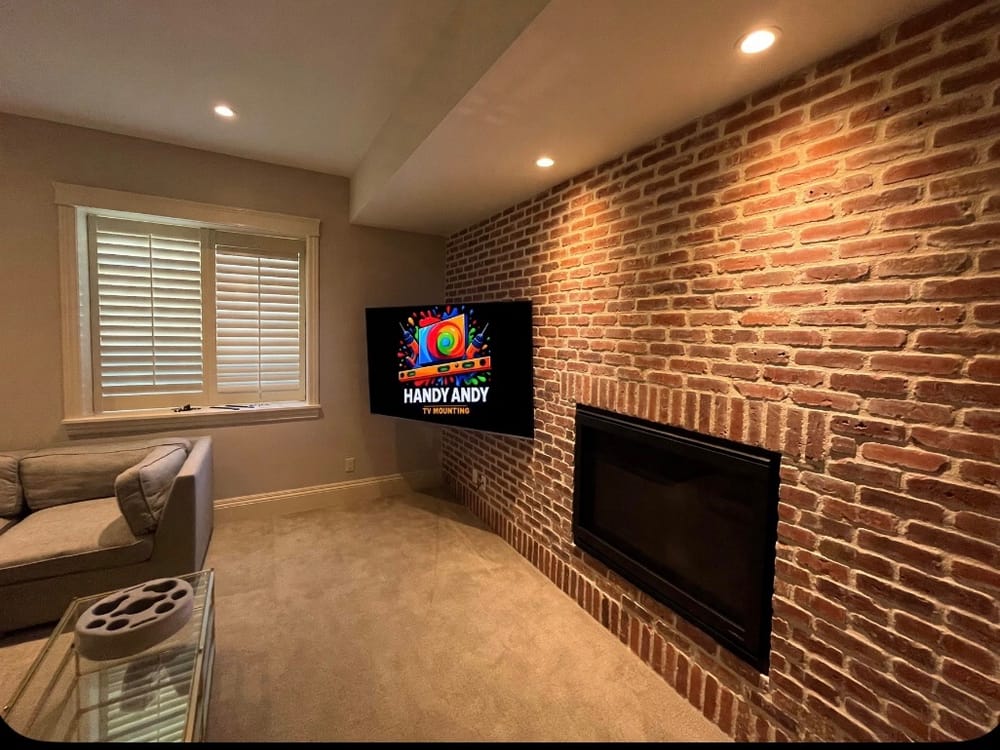 TV mounted on a wall beside an exposed brick fireplace wall with plantation shutters, displaying the Handy Andy logo