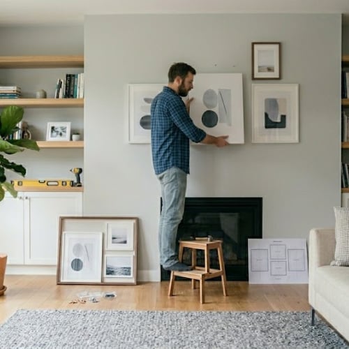 Man in plaid shirt hanging framed artwork on living room wall above fireplace, arranging home decor