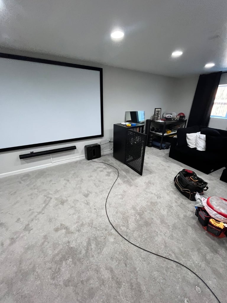 Home theater room with large projection screen, black soundbar, desk with equipment, and cable on concrete floor
