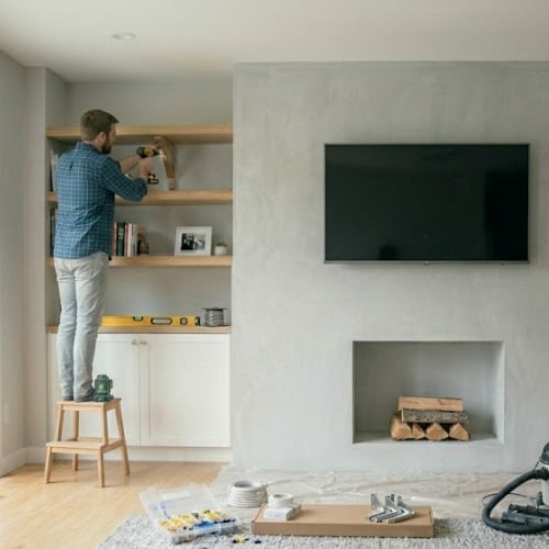 Man standing on wooden stool arranging items on built-in wall shelves in modern living room with fireplace and TV