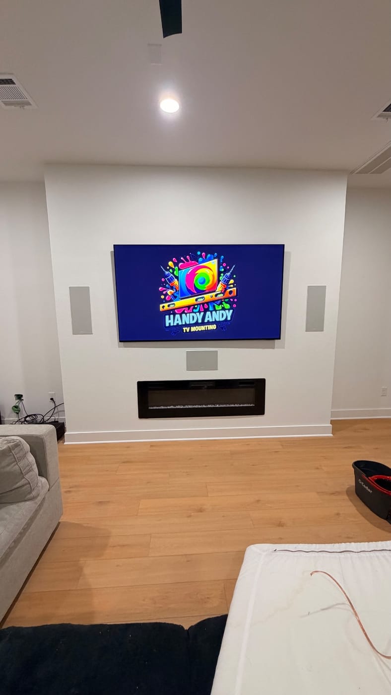 TV mounted above a linear electric fireplace with in-wall speakers on each side, displaying the Handy Andy logo