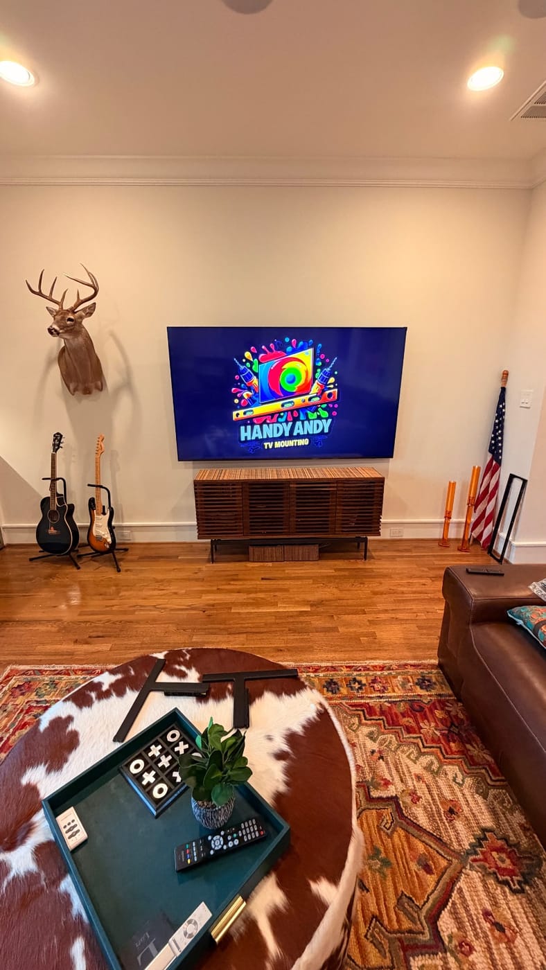 TV mounted above a wood media console in a living room with a mounted deer head and guitars, displaying the Handy Andy logo