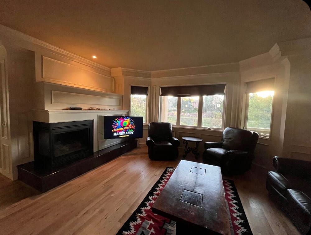 TV mounted above a fireplace in a living room with bay windows and leather chairs, displaying the Handy Andy logo