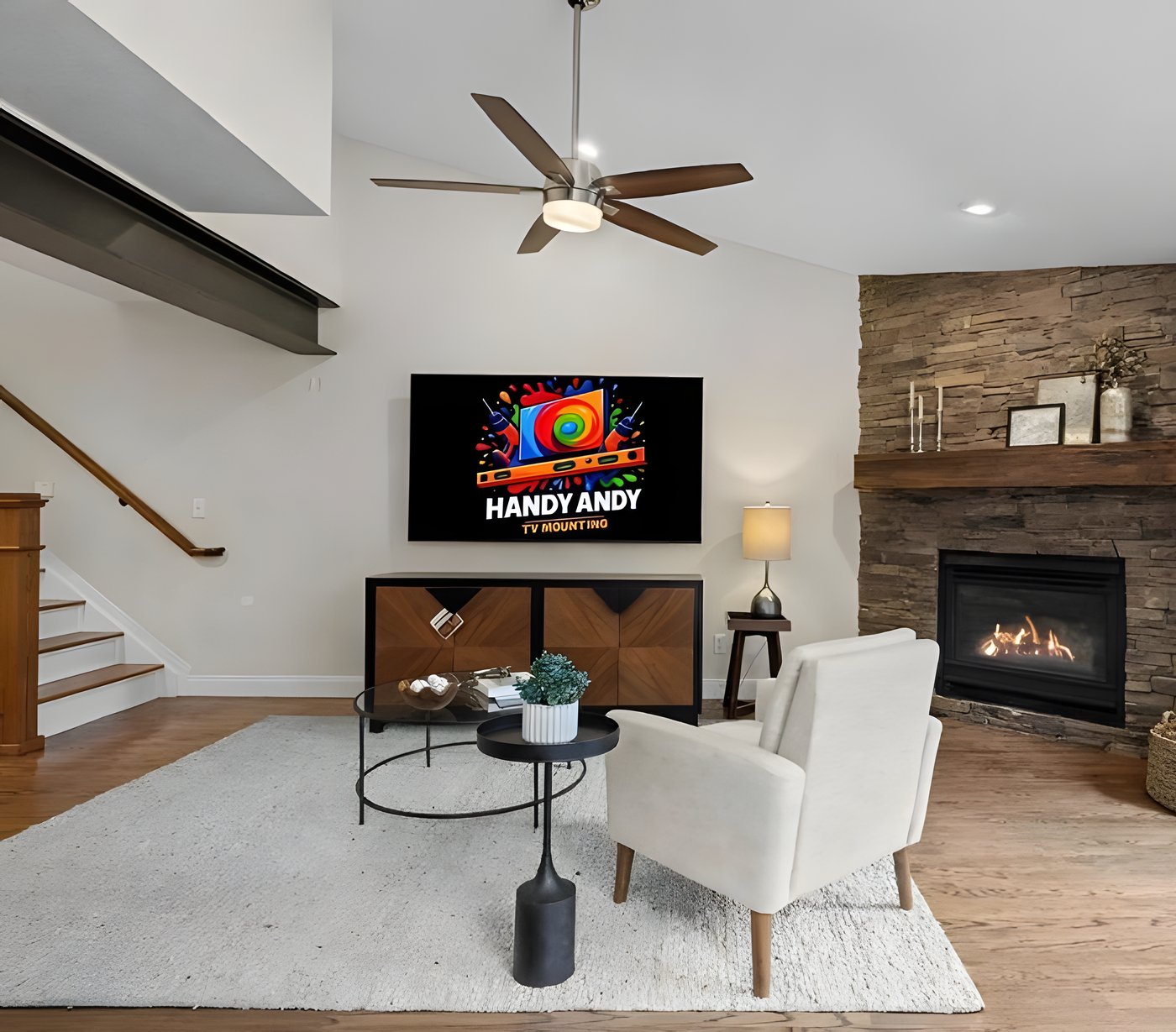 TV mounted above a wood media console in a living room with a stacked-stone corner fireplace, showing the Handy Andy logo
