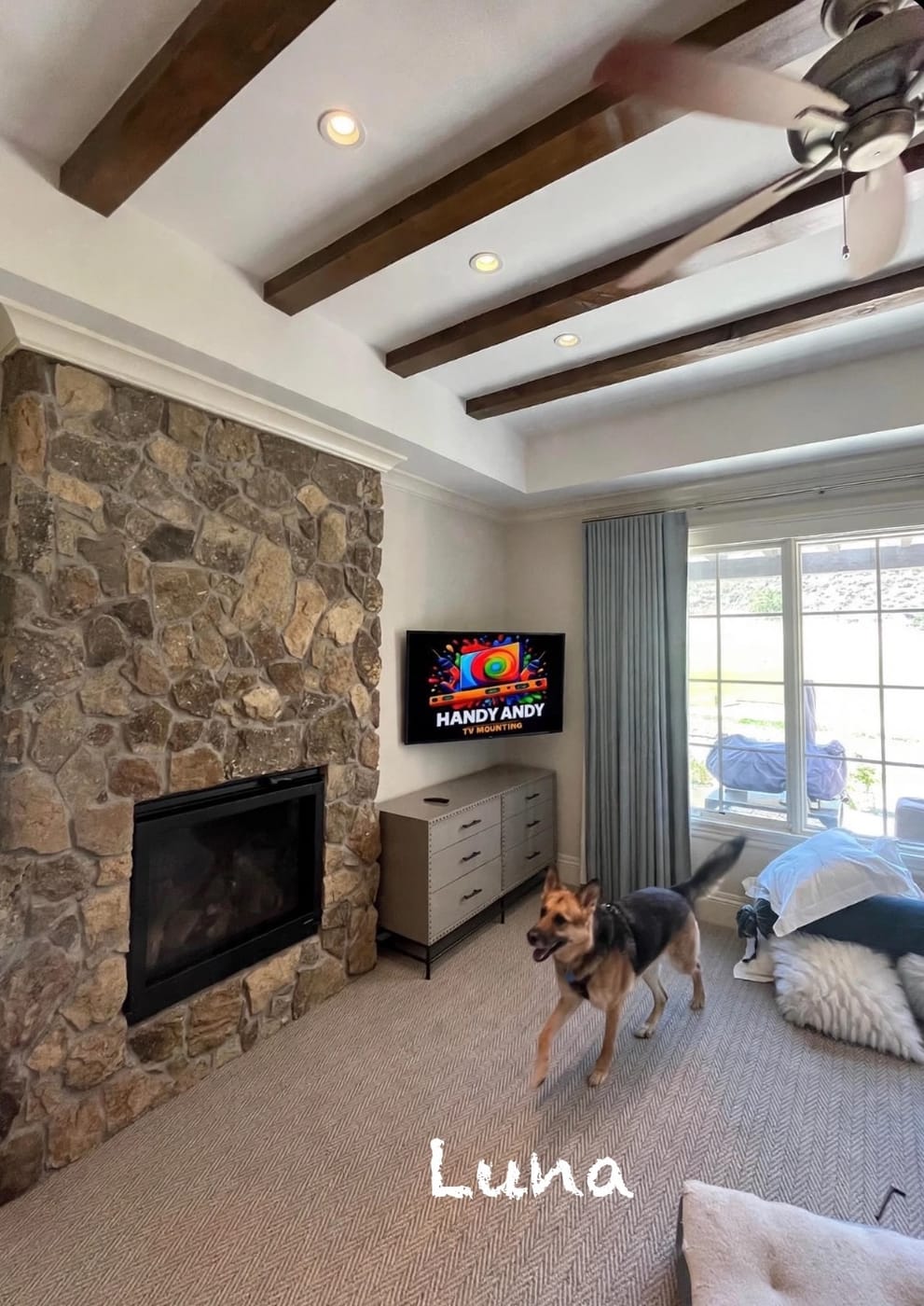 TV mounted beside a stone fireplace wall under exposed ceiling beams, displaying the Handy Andy logo, with a dog in the room
