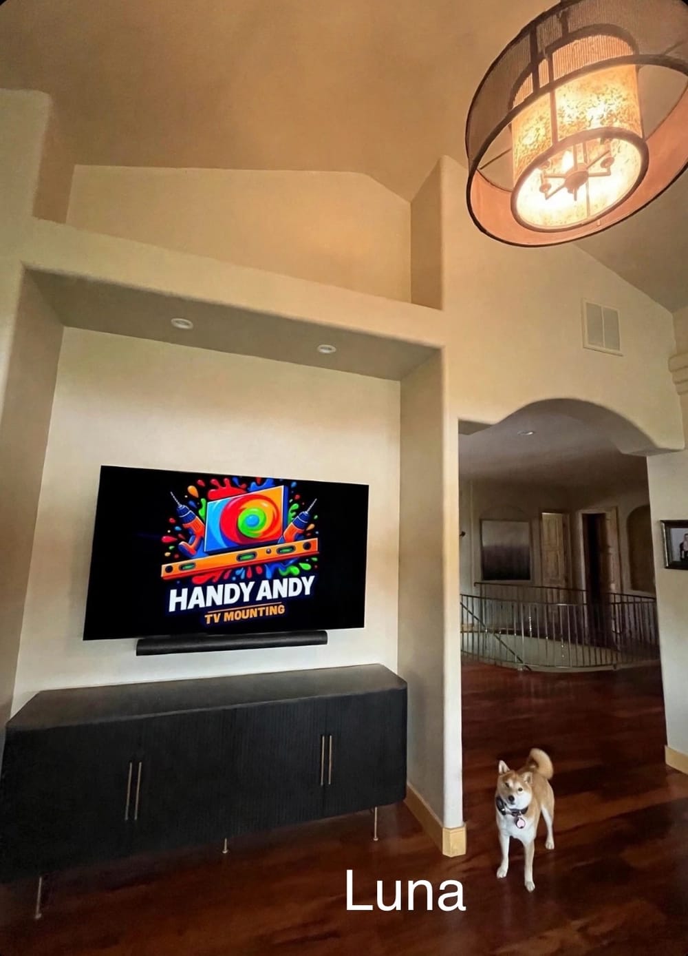 TV mounted above a dark console cabinet in an arched hallway living room, displaying the Handy Andy logo, with a small dog on the wood floor