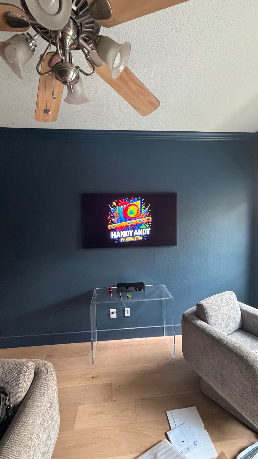 TV mounted on dark accent wall above a clear acrylic console table displaying the Handy Andy logo