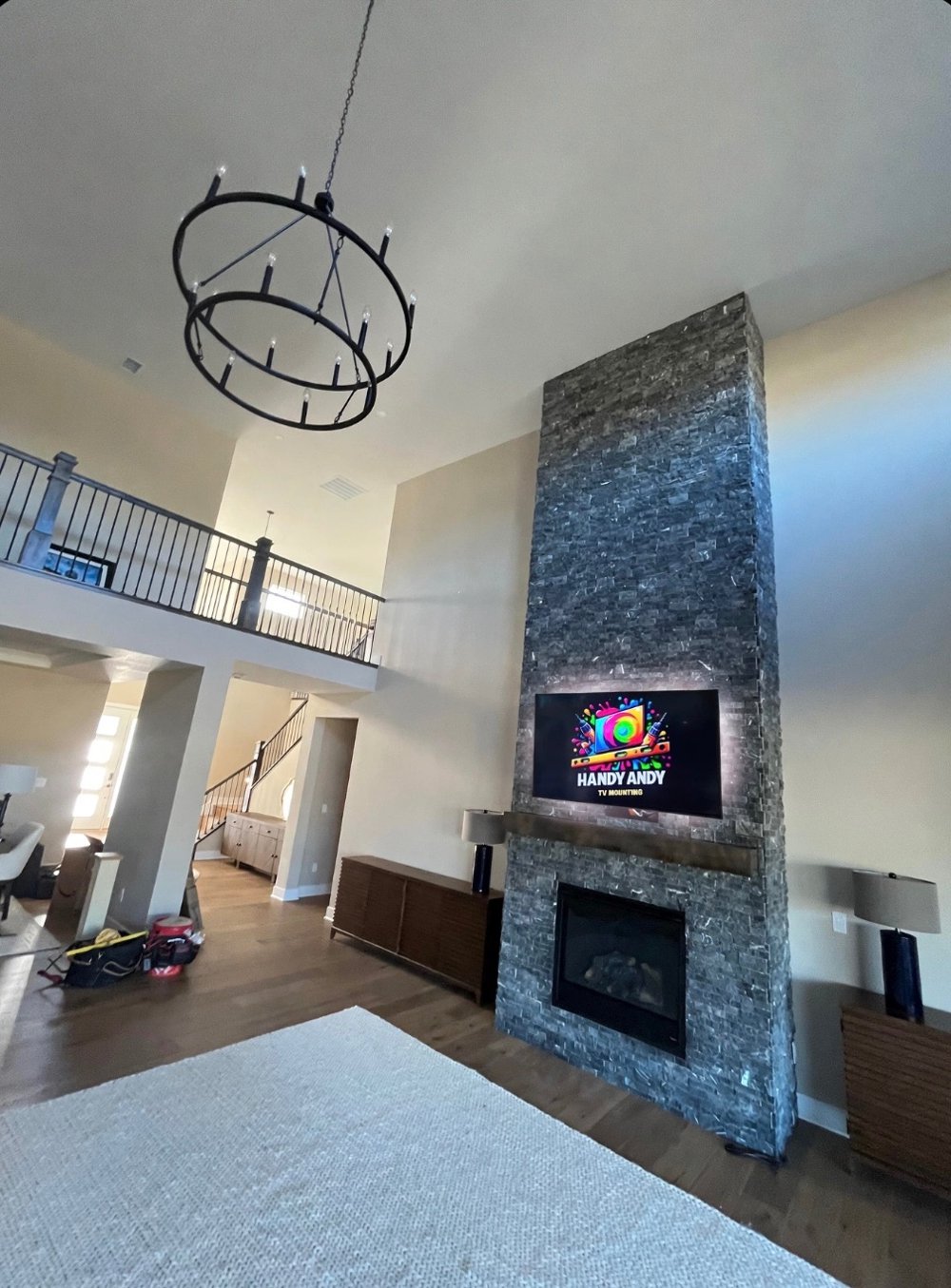 TV with LED backlighting mounted above a gas fireplace on a tall gray stacked stone wall under a two-story ceiling