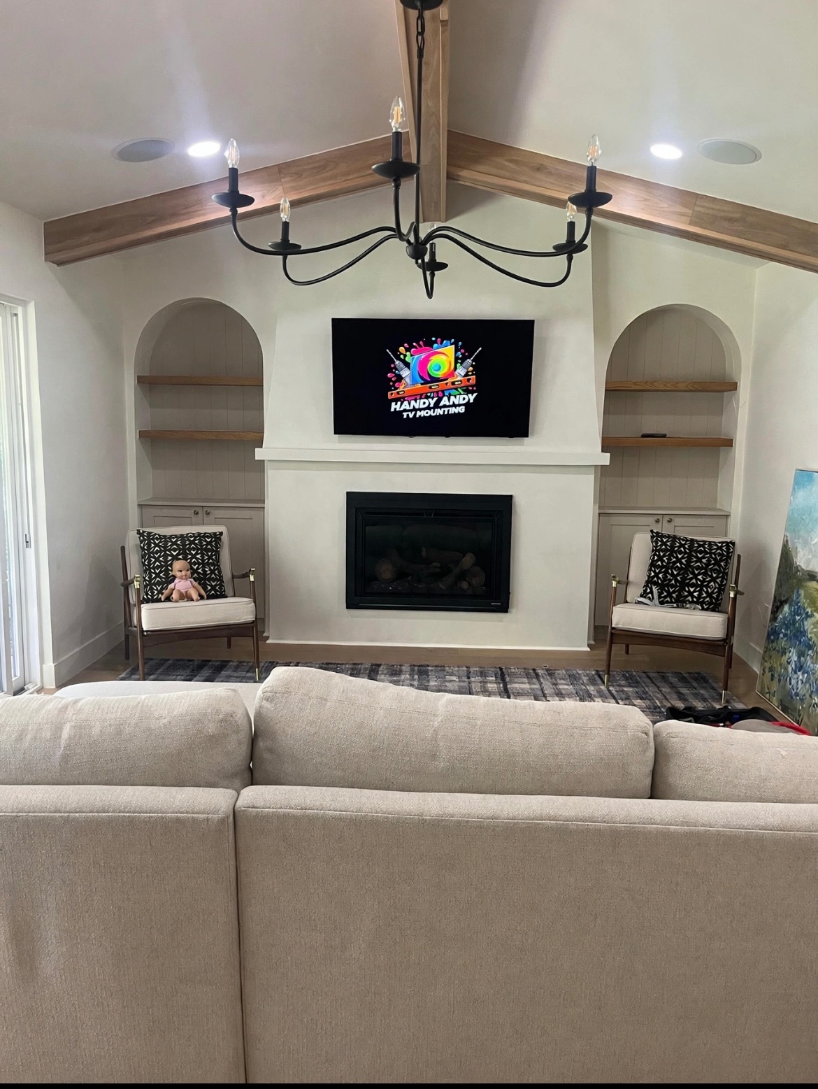 TV mounted above a fireplace with vaulted ceiling, wood beams, arched built-in shelving and a chandelier displaying the Handy Andy logo