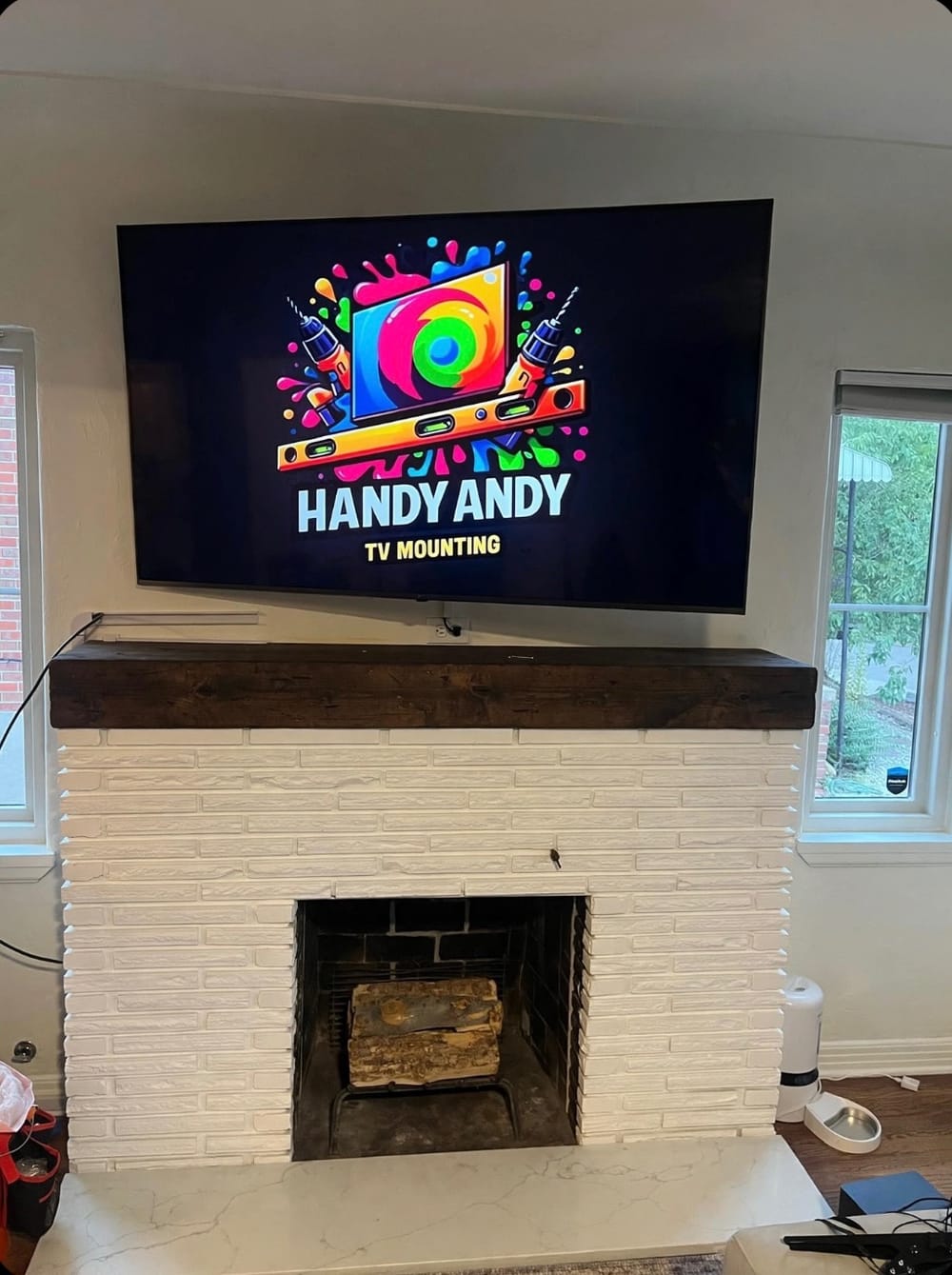 Large TV mounted above a white brick fireplace with a dark wood mantel, displaying the Handy Andy logo