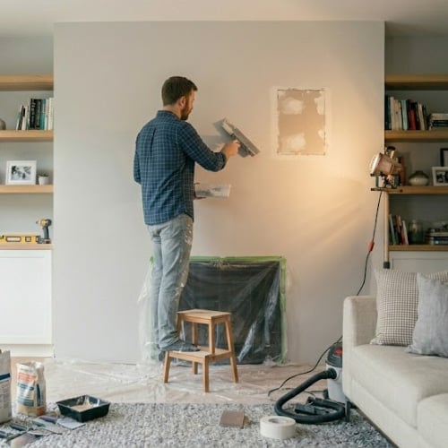 Man in blue plaid shirt using putty knife on drywall, standing on wooden stool during home renovation with furniture and tools visible