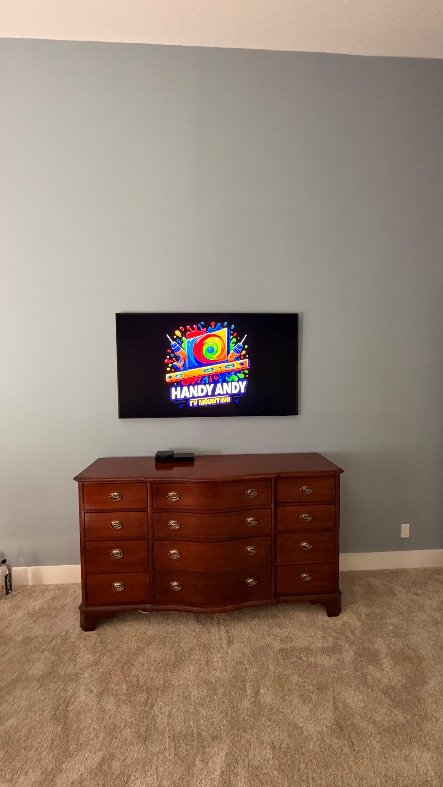 TV mounted above a wood dresser in a bedroom displaying the Handy Andy logo