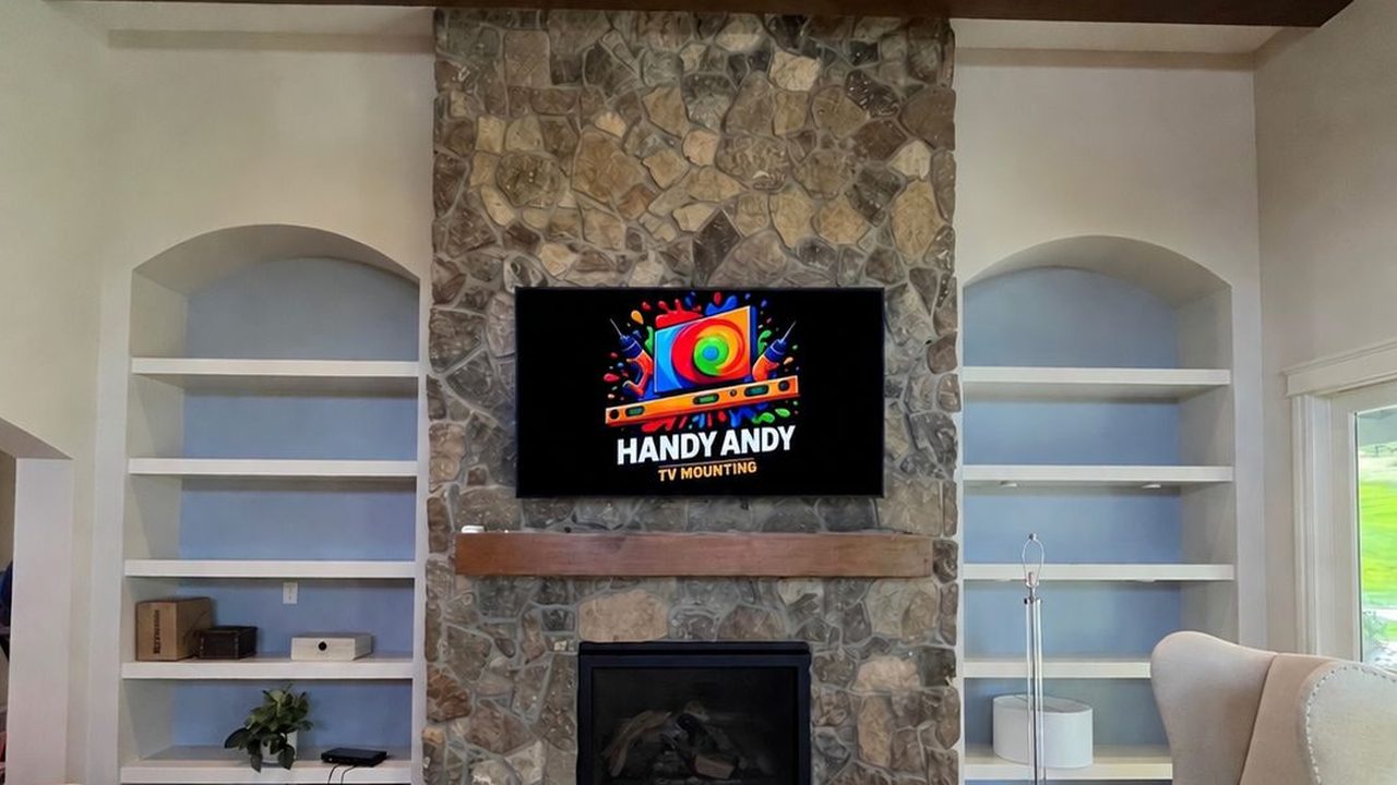 TV mounted level and centered on a stacked-stone fireplace above a wood mantel, between built-in shelves, showing the Handy Andy TV Mounting logo