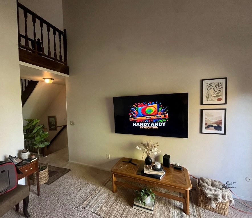 TV mounted on a tall living room wall under a loft beside two framed prints, showing the Handy Andy logo