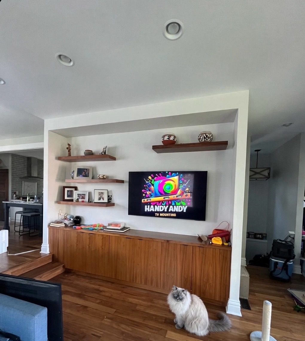 TV mounted in a white wall niche between floating walnut shelves above a walnut built-in cabinet, showing the Handy Andy logo