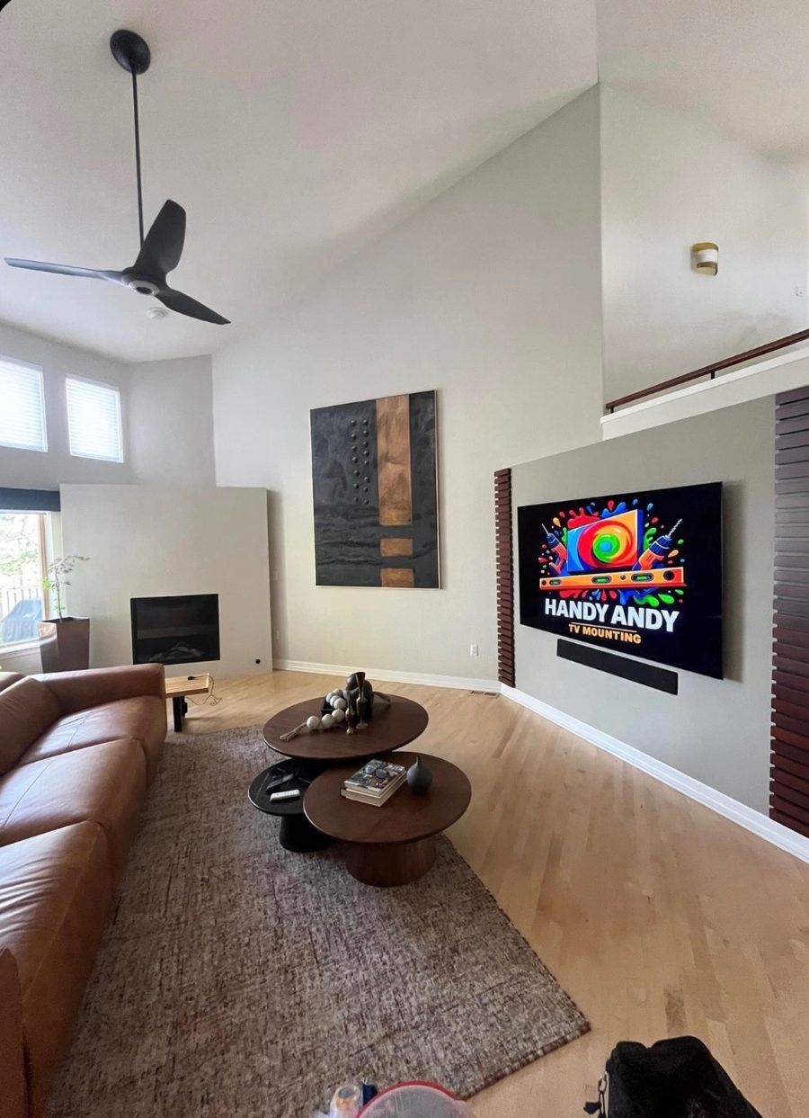 TV mounted on a gray accent wall framed by dark wood slats, soundbar below, in a vaulted living room with a ceiling fan, showing the Handy Andy logo