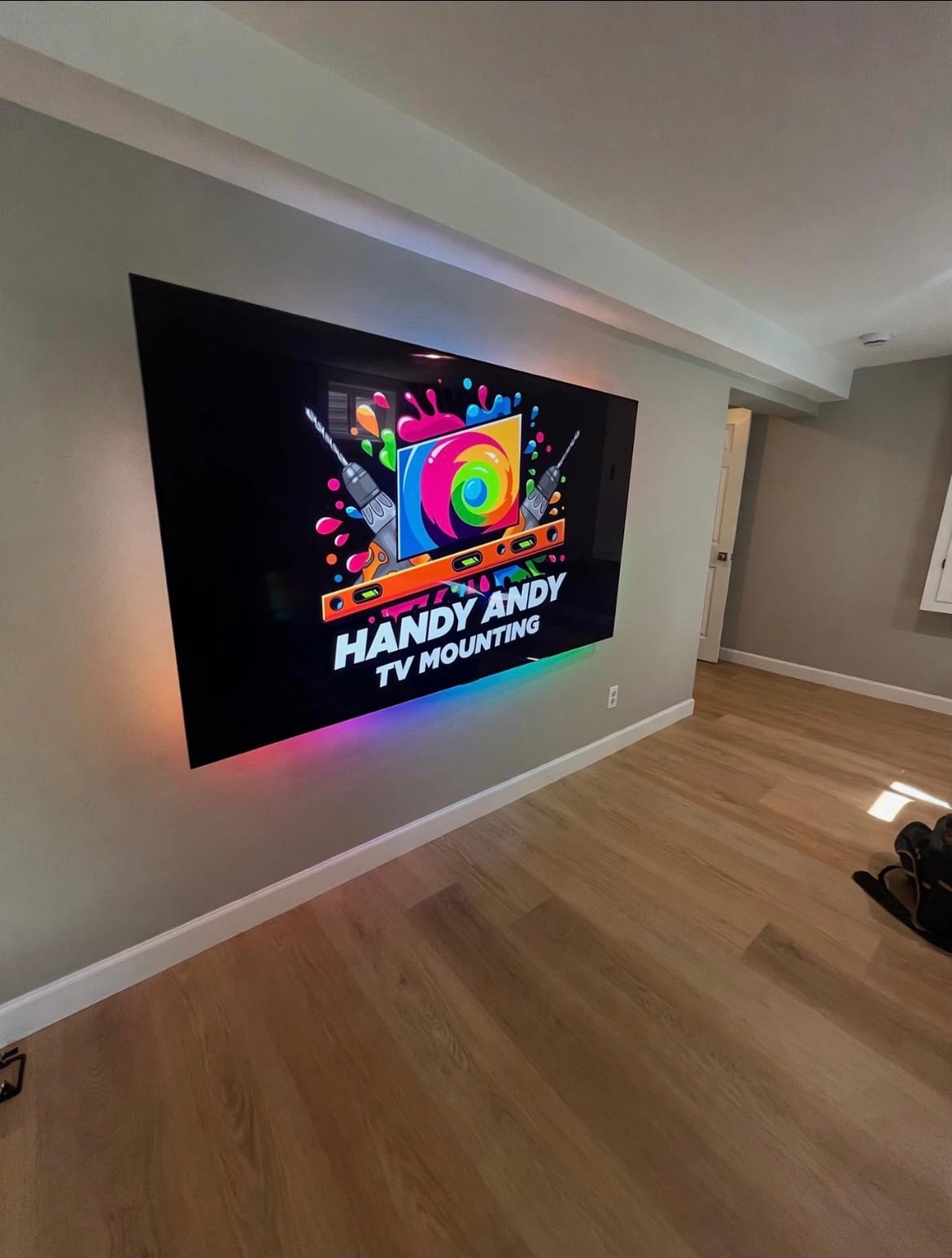 TV wall-mounted in a hallway with hardwood floors, screen displaying the Handy Andy TV Mounting logo