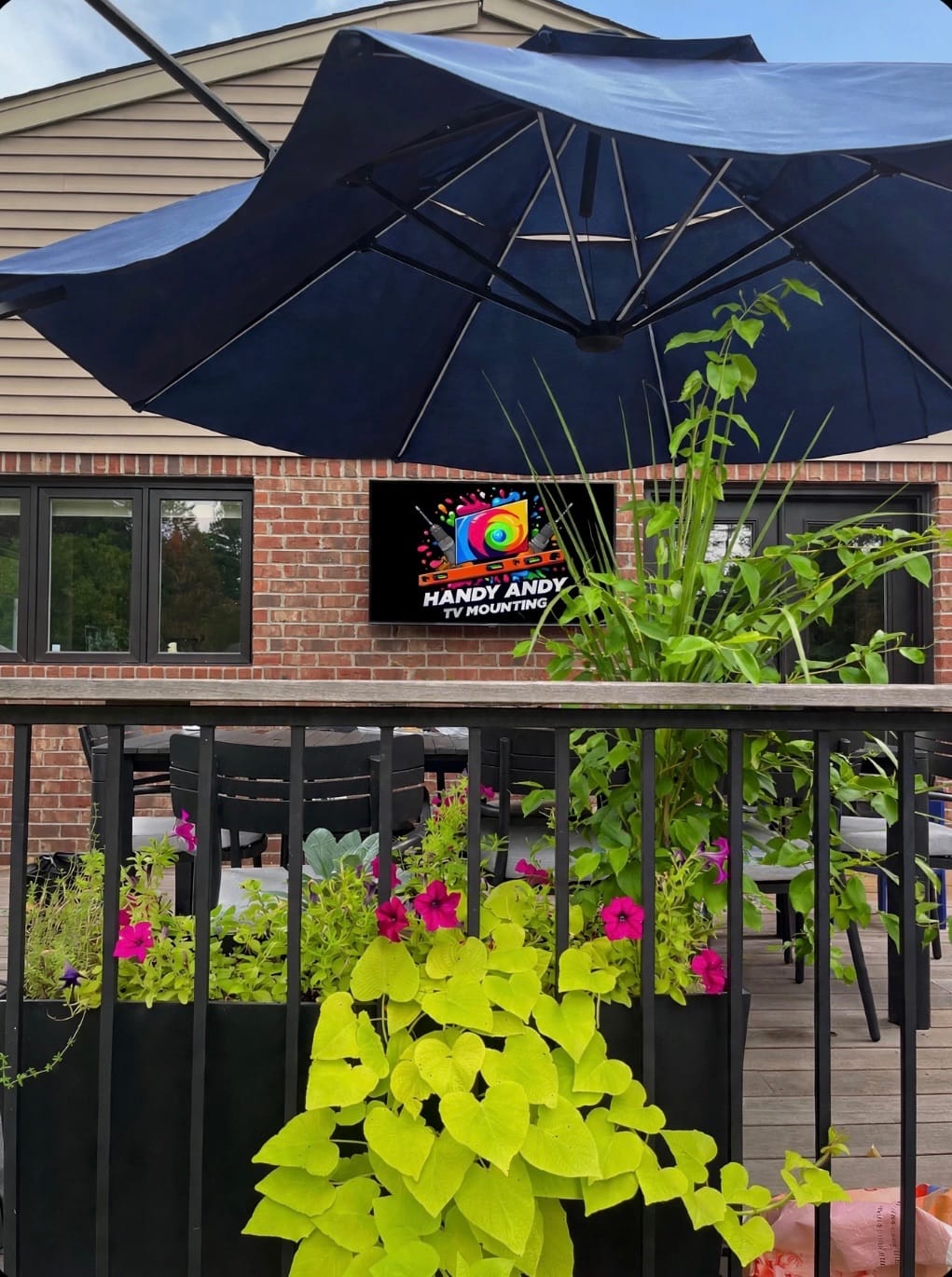 Outdoor TV mounted under a patio umbrella surrounded by hanging plants, screen displaying the Handy Andy TV Mounting logo